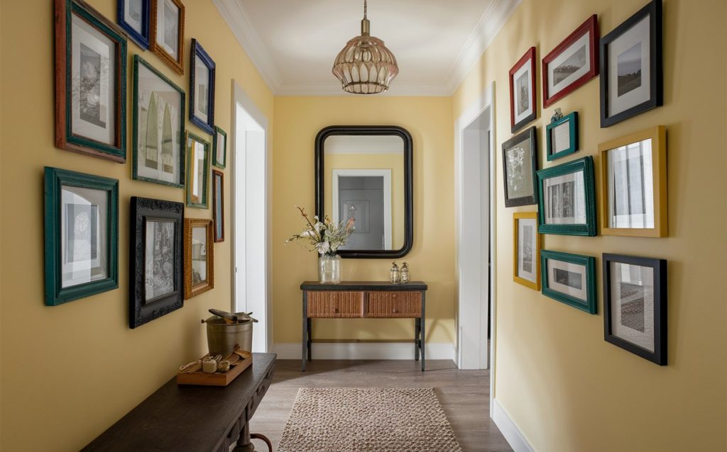 A narrow hallway with pastel yellow walls and a variety of multi-colored picture frames hanging on them.