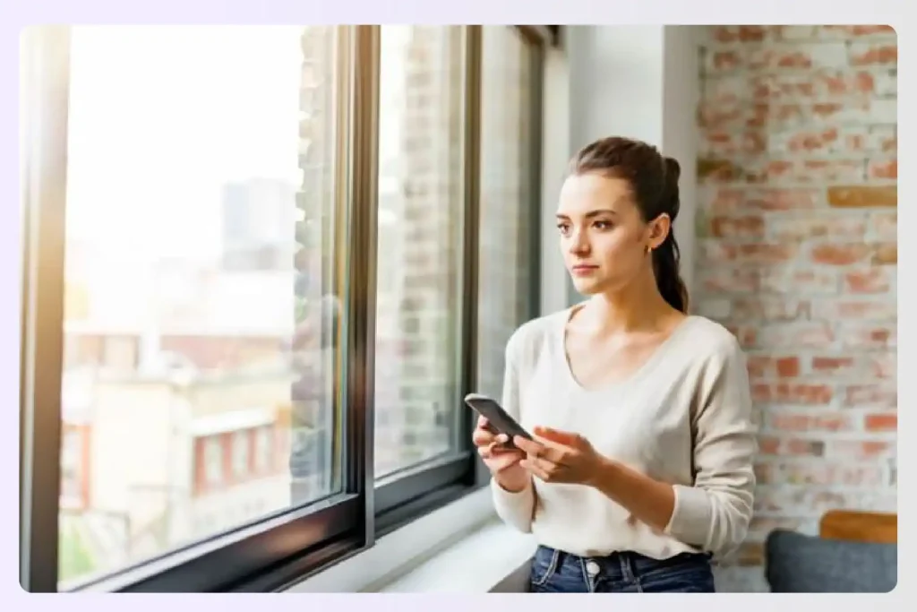 Woman looking out a window while holding her smartphone